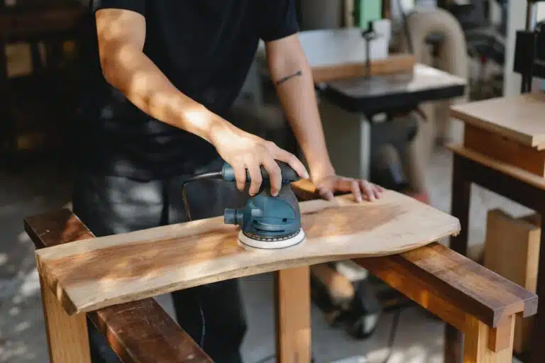 Artesanato em madeira: 15 ideias para vender Mãos de um homem de camiseta preta manipulando uma lixadeira numa peça de madeira.